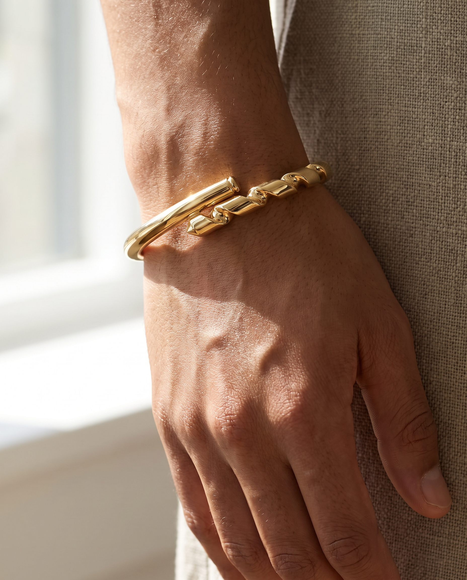 A close-up of a person's hand and wrist wearing a chunky gold chain-link bangle bracelet, set against a neutral textured fabric background.